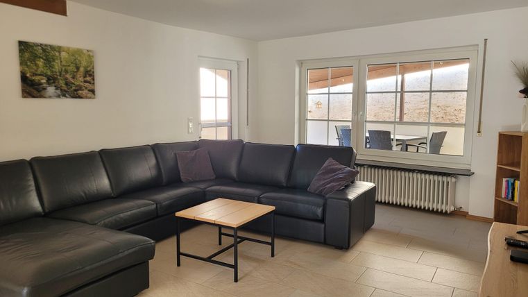 Ein modernes Wohnzimmer mit einer großen schwarzen Couch und einem Holztisch. Durch die Fenster ist eine Aussicht auf einen Balkon zu sehen.