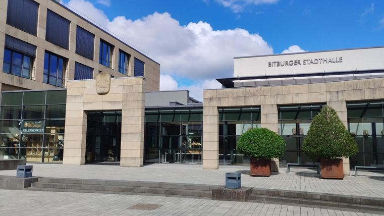 Eine moderne Eingangshalle mit großen Fenstern und einer klaren Fassade. Im Hintergrund sieht man einen blauen Himmel mit einigen Wolken.