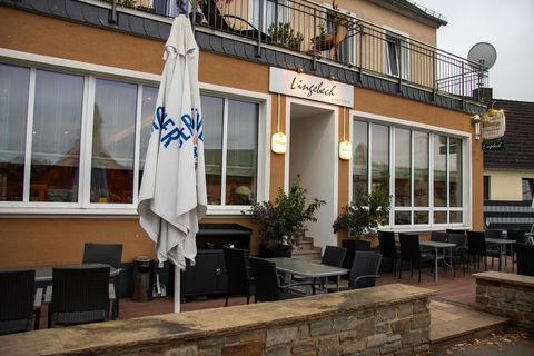 Ein gemütliches Restaurant mit einer Terrasse und mehreren Tischen. Die Fassade ist hell erleuchtet, und ein Sonnenschirm steht vor dem Eingang.