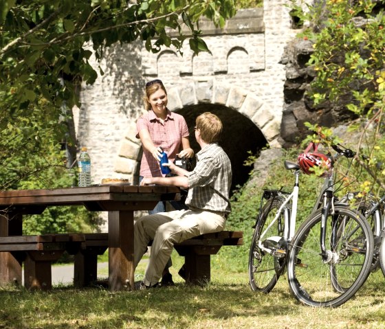 Zwei Personen sitzen an einem Picknicktisch im Gr&uuml;nen, Fahrr&auml;der und eine Steinbr&uuml;cke im Hintergrund. Sie genie&szlig;en eine Pause am Enz-Radweg., &copy; Eifel Tourismus GmbH/D. Ketz