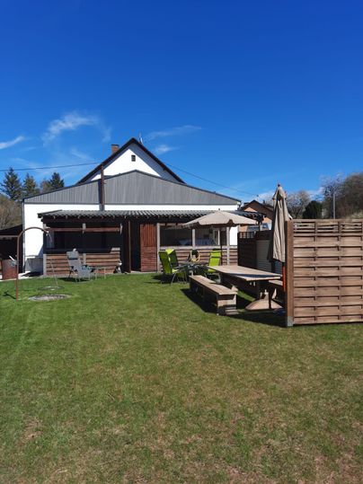 Ein einladender Garten mit einem Haus im Hintergrund. Es gibt Sitzgelegenheiten und Sonnenschirme unter einem klaren blauen Himmel.