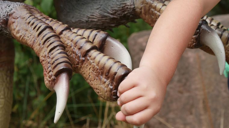 Een kind houdt de klauwen van een dinosaurussculptuur vast. De scène toont een interactie tussen het kind en de sculptuur.
