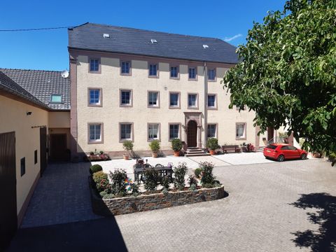 Ein großes, helles Gebäude mit einem geräumigen Innenhof. Im Vordergrund sind Blumenbeete und ein rotes Auto zu sehen.