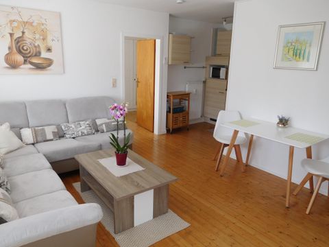 Een lichte woonkamer met een gezellige bank, een tafel en een kleine eettafel. Een orchidee in een pot zorgt voor een frisse accent in de ruimte.