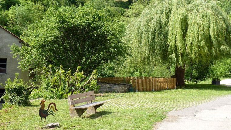 A tranquil rural scene with a wooden bench and a large willow tree. In the foreground, there is a chicken symbol on the lawn.