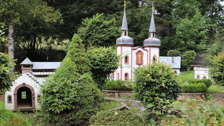 Eine kleine Gartenlandschaft mit zwei Türmen und einer bunten Kirche. Umgeben von üppigem Grün und Hecken.