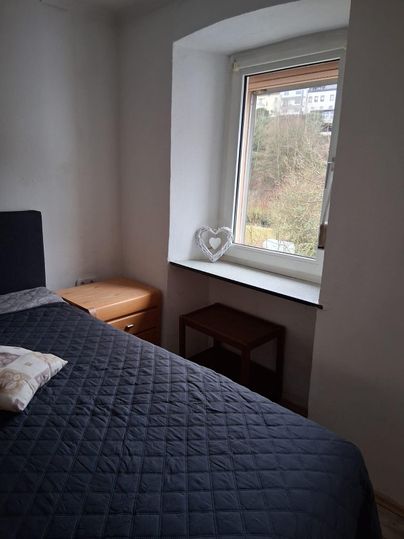 Une chambre simple avec un lit et une fenêtre. Dehors, un paysage vert est visible.