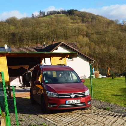 A red Volkswagen is parked in front of a house in a rural setting. In the background, gentle hills and trees can be seen.