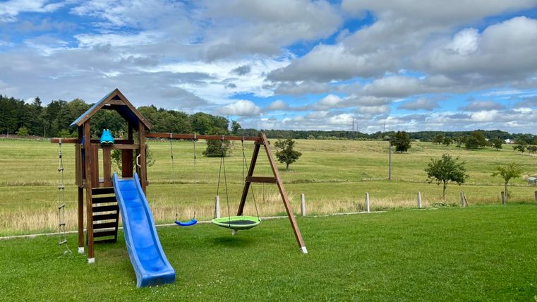 Ein Spielplatz mit einer Rutsche und einer Schaukel steht auf einer grünen Wiese. Im Hintergrund sind Bäume und ein klarer Himmel zu sehen.
