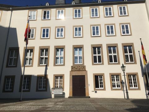 Een historisch gebouw met meerdere ramen en twee vlaggen. De gevel is helder en duidelijk, omringd door een plein.