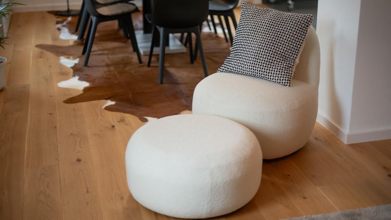 Une pièce moderne avec une chaise confortable et un pouf rond dans des couleurs claires. Le sol est en bois et il y a une table à manger à l'arrière-plan.