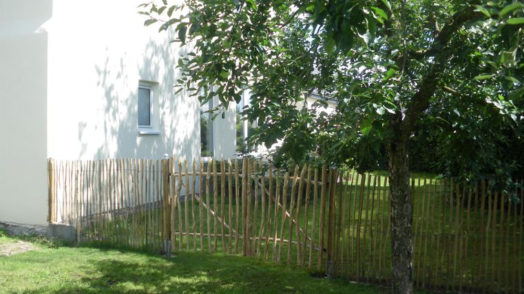 Ein Haus mit einem Garten, umgeben von einem Holzlattenzaun. Neben dem Haus steht ein grüner Baum im Schatten.