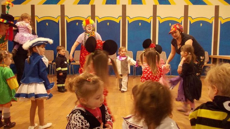 Eine fröhliche Kindermaskerade mit vielen verkleideten Kindern in bunten Kostümen. Im Hintergrund spielen Clowns und Erwachsene mit, während die Kinder tanzen und lachen.