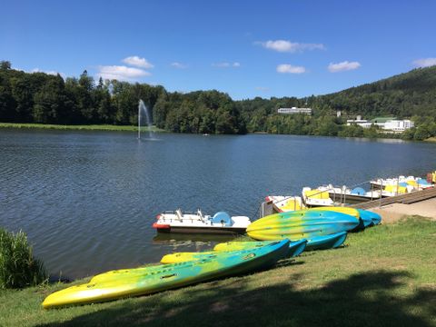 Ein ruhiger See mit bunten Kajaks und Tretbooten am Ufer. Im Hintergrund sind Bäume und ein Freizeitbereich zu sehen.