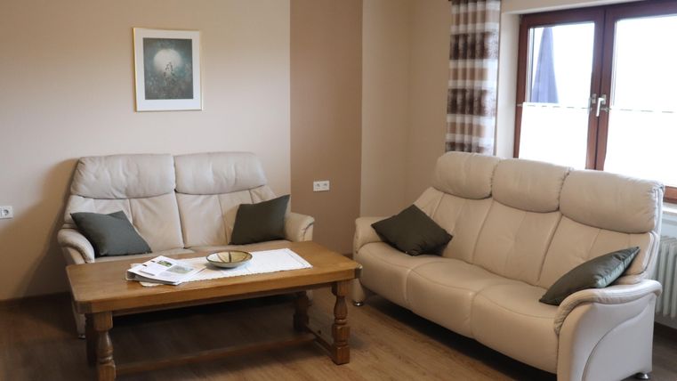 Un salon confortable avec deux canapés beiges et une table basse en bois. Un tableau est accroché au mur et la lumière passe à travers les fenêtres.