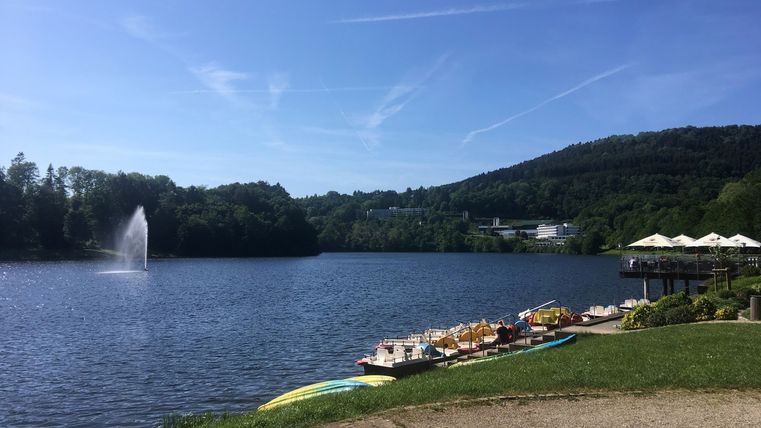 Bitburger Stausee mit Tretbooten und Springbrunnen, umgeben von Bäumen und Hügeln.