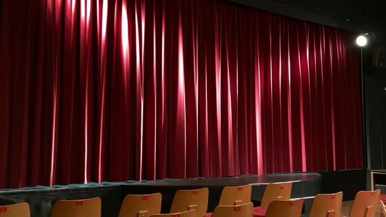 Ein Theatersaal mit roten Vorhängen und leeren Holzbänken. Das Licht fällt sanft auf die Vorhänge, während die Bühne wartet.