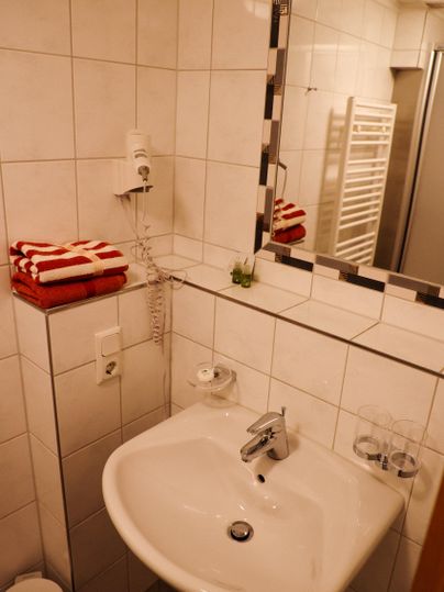 Une petite salle de bains avec des carreaux blancs et un lavabo. Des serviettes rouges et un mur miroir complètent l'équipement.