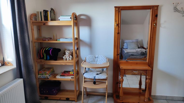 Ein Zimmer mit einem Holzregal und einem Schminktisch. Im Regal sind Bücher und ein Stofftier, daneben steht ein Hochstuhl.