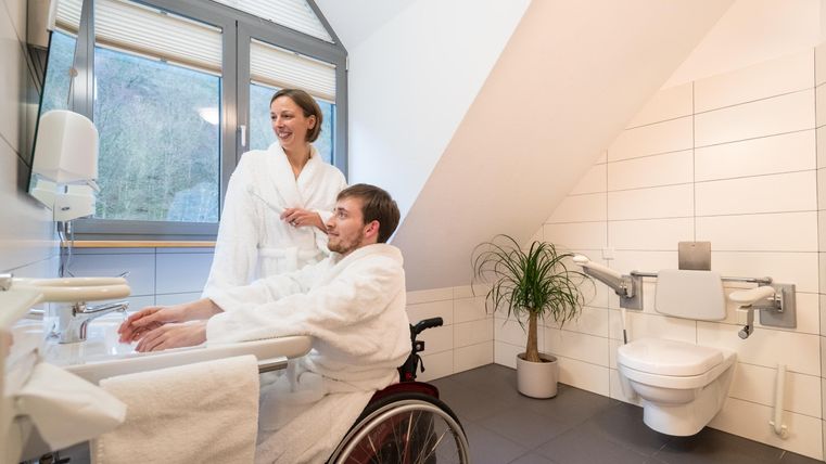 Een moderne, toegankelijke badkamer met een rolstoelvriendelijke wastafel en toilet. Twee personen in badjassen staan in de ruimte en zien er ontspannen uit.