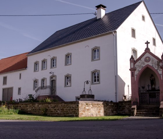 Ein altes Bauernhaus mit wei&szlig;er Fassade und rotem Dach, daneben eine kleine Kapelle mit Kreuz. Der Himmel ist blau und klar., &copy; Tourist-Information Bitburger Land