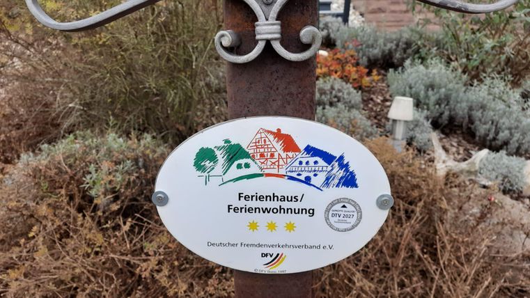 Ein Schild, das auf ein Ferienhaus oder eine Ferienwohnung hinweist. Es zeigt Symbole und Auszeichnungen für die Unterkunft.