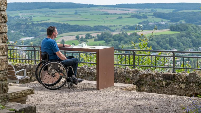 Ein Mann im Rollstuhl sitzt an einem Tisch mit Blick auf die Landschaft. Die Umgebung ist grün und hügelig, und der Himmel ist teilweise bewölkt.