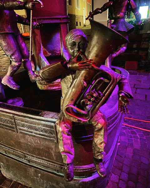 Eine metallene Statue eines Musikers, der eine Tuba spielt. Die Figur sitzt auf einer Brunnenmauer und ist in sanftes Licht getaucht.