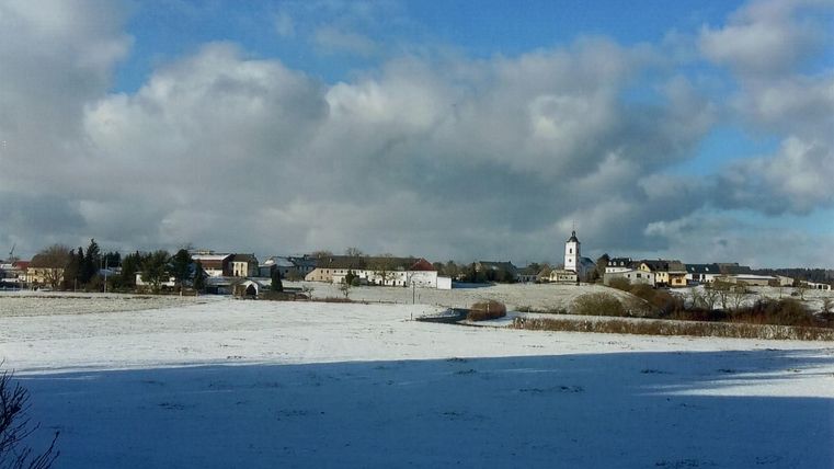 A winter landscape with a snow-covered field and a small village in the background. The sky is partly cloudy and radiates in various shades of blue.