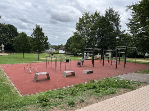 A playground with fitness equipment on slip-resistant ground. In the background, green trees are visible and the sky is cloudy.
