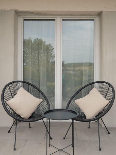A cozy balcony with two black armchairs and a small table. The armchairs are adorned with light-colored cushions, and in the background, you can see windows with curtains.