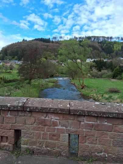 Un paysage pittoresque avec une rivière qui coule et une verdure luxuriante. À l'arrière-plan, on voit des collines douces et des arbres.