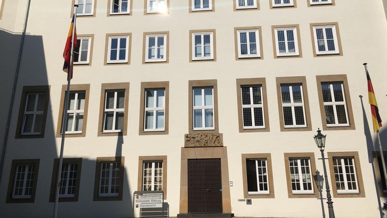 Een historisch gebouw met meerdere ramen en twee vlaggen. De gevel is helder en duidelijk, omringd door een plein.