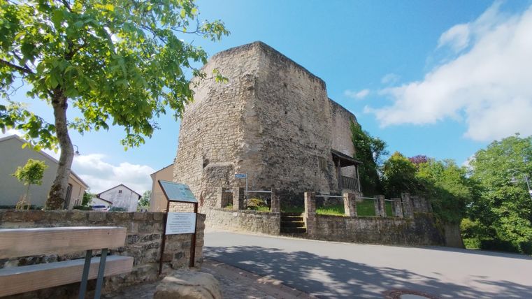 Ein alter Steinturm steht an einer Straße, umgeben von Bäumen. Der blaue Himmel sorgt für eine freundliche Atmosphäre.