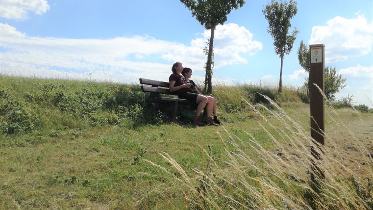 Zwei Personen sitzen auf einer Bank in einer grünen Landschaft mit Bäumen und blauem Himmel.