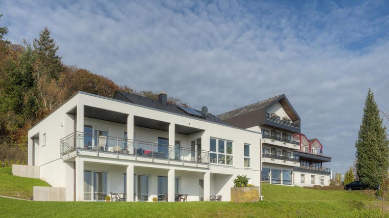 Moderne Gebäude auf einer grünen Wiese, umgeben von Bäumen. Der Himmel ist teilweise bewölkt.