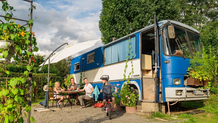 Een blauwe bus staat in een groene omgeving. Een gezin zit aan de tafel in de openlucht en geniet van de tijd samen.