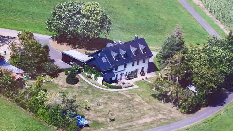 Une grande maison moderne entourée de prairies vertes et d'arbres. L'allée mène doucement au bâtiment, qui est entouré d'un terrain bien entretenu.