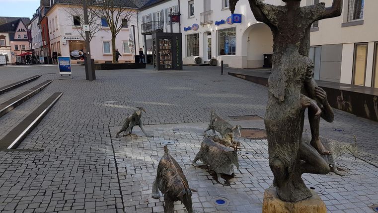 Eine belebte Platz mit einer Statue von Menschen und Tieren. Umgeben von historischen Gebäuden und gepflasterten Wegen.