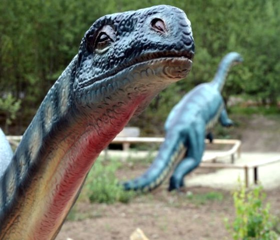 A dinosaur model in the Dinopark, surrounded by green vegetation. Another dinosaur model can be seen in the background.