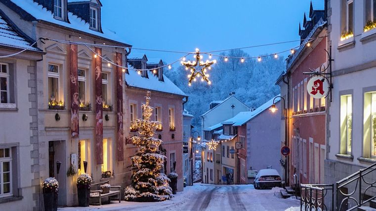 Eine schneebedeckte Straße mit festlich geschmückten Häusern. Lichter und ein Weihnachtsbaum erzeugen eine gemütliche Atmosphäre in der Dämmerung.