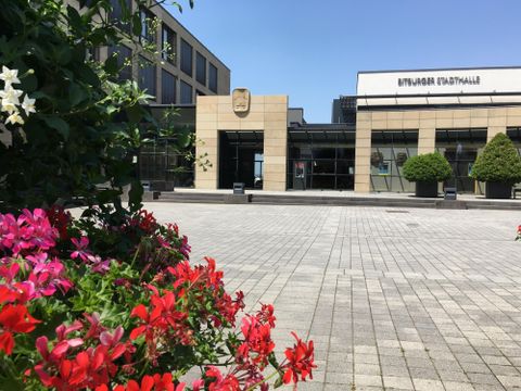 Ein modernes Gebäude mit einer großzügigen Fassade und großen Fenstern. Vor dem Gebäude gibt es farbenfrohe Blumenbeete und eine gepflasterte Fläche.
