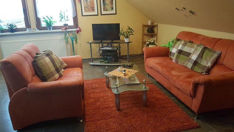 Un salon confortable avec deux canapés orange et une table basse en verre. À l'arrière-plan, il y a des plantes et une télévision sur une étagère.