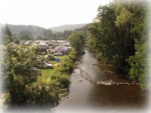 Un ruisseau tranquille coule à côté d'un terrain de camping, entouré d'arbres. En arrière-plan, des tentes et des caravanes sont visibles.