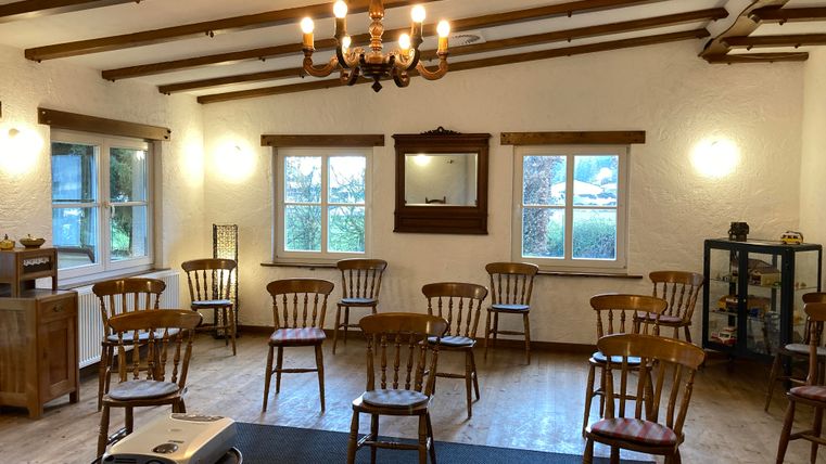 Une pièce confortable avec des chaises en bois et une table simple. Elle a des fenêtres donnant sur l'extérieur et une atmosphère chaleureusement éclairée.