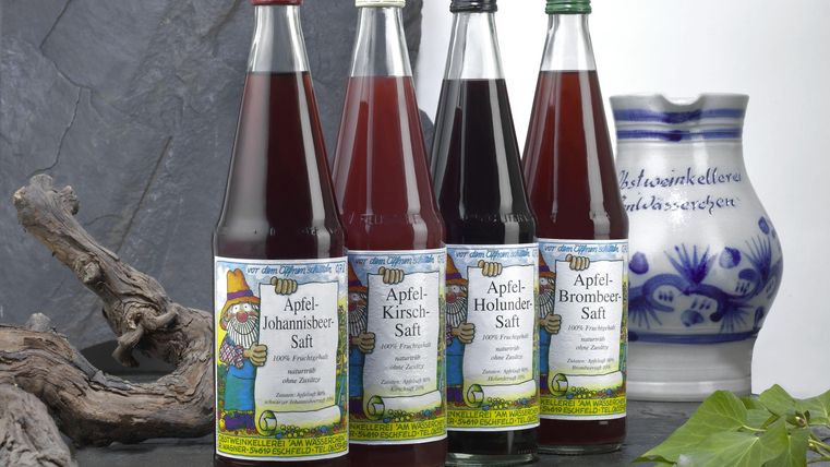 A selection of four bottles of apple juice in different varieties. In the background, there is a decorative ceramic carafe.