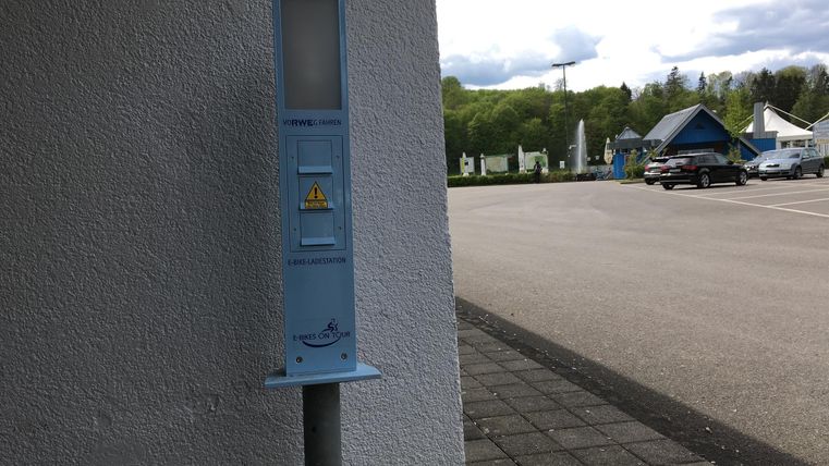 Ein blauer Parkscheinautomat steht an einer Wand. Im Hintergrund ist ein Parkplatz mit Fahrzeugen und umgebender Natur sichtbar.