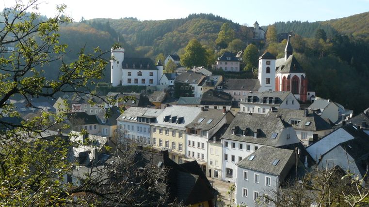 Vue de la ville de Neuerburg avec des bâtiments historiques et une église, entourée de collines boisées.