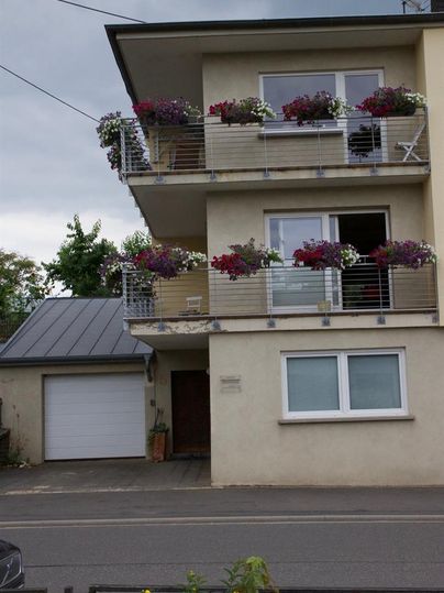 Ein modernes Gebäude mit einem Balkon voller blühender Pflanzen. Die Fassade ist hell und freundlich, und es gibt eine Garage auf der linken Seite.