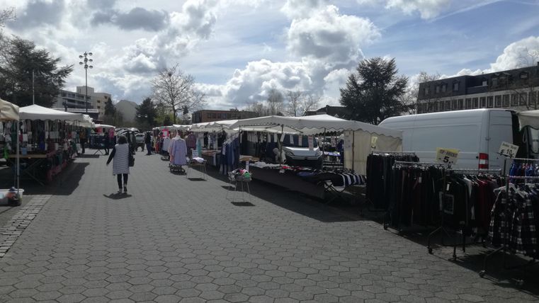 Ein belebter Markt mit Verkaufsständen und zahlreichen Waren. Der Himmel ist bewölkt, und viele Menschen schlendern umher.
